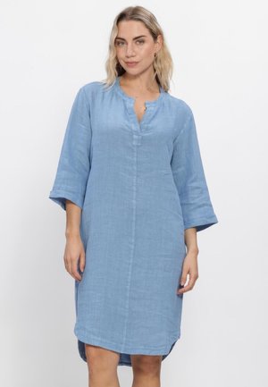 Vrouw draagt een lichtblauwe, knielange linnen jurk met driekwartsmouwen en een kleine V-hals, staande tegen een witte achtergrond.