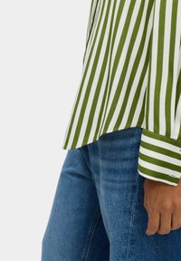 Camicia a righe verdi e bianche con maniche lunghe, fessura laterale e vestibilità rilassata, abbinata a jeans blu. Il tessuto appare liscio con una leggera lucentezza.