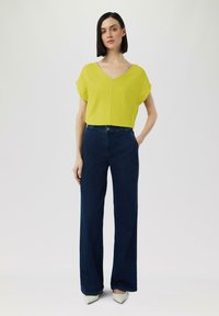 Jasnozielona bluzka z krótkim rękawem i dekoltem w serek, połączona z wysokimi, ciemnoniebieskimi spodniami typu wide-leg oraz jasnymi, spiczastymi butami.