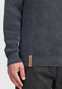 Pull en maille bleu marine texturé avec des poignets et un ourlet côtelés, arborant un motif tissé. Inclut une étiquette en cuir avec "JEANSCODE" sur le côté.