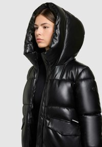 Giubbotto impermeabile nero con una texture lucida e liscia, cappuccio oversize e grandi tasche frontali. Presenta una chiusura con cerniera e un dettaglio con bottone sul maniche.