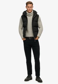 Schwarze Pufferweste mit Kapuze über einem hellgrauen, gerippten Rollkragenpullover, kombiniert mit schwarzen Hosen und schwarzen Schnürschuhen.