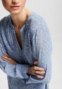 Blusa azzurro chiaro con un motivo a pois in bianco e blu scuro, con scollatura con bottoni e maniche lunghe leggermente gonfie.