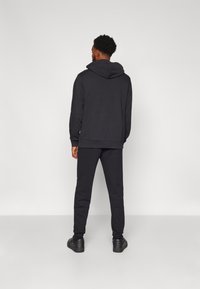 Felpa nera con cappuccio regolabile, abbinata a pantaloni della tuta affusolati e scarpe da ginnastica nere. Il tessuto sembra morbido e strutturato.