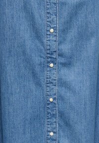 Chemise en denim de couleur bleu clair, coupe droite, avec une patte de boutonnage avant et des boutons blancs. Texture lisse avec un délavage minimal.