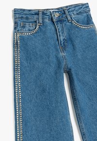 Jeans de mezclilla azul con un diseño de pierna recta, que presentan tachuelas plateadas a lo largo de la costura lateral y bolsillos tradicionales, terminados con costuras naranjas.
