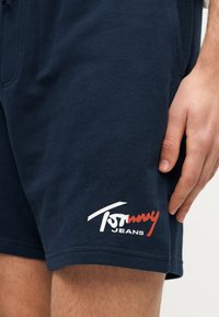 Κοντινό πλάνο στο μηρό ενός ατόμου που φορά σκούρα μπλε σορτς με λευκό και κόκκινο λογότυπο "Tommy Jeans" στο κάτω μέρος του ποδιού.