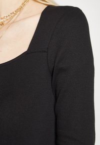 Top preto canelado de manga longa com decote quadrado e silhueta ajustada, apresentando um tecido texturizado e costuras dos ombros ligeiramente bufantes.