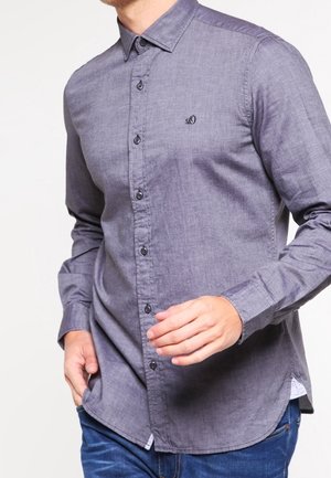 Homme portant une chemise grise à manches longues boutonnée avec un petit logo brodé sur la poitrine et un jean bleu.
