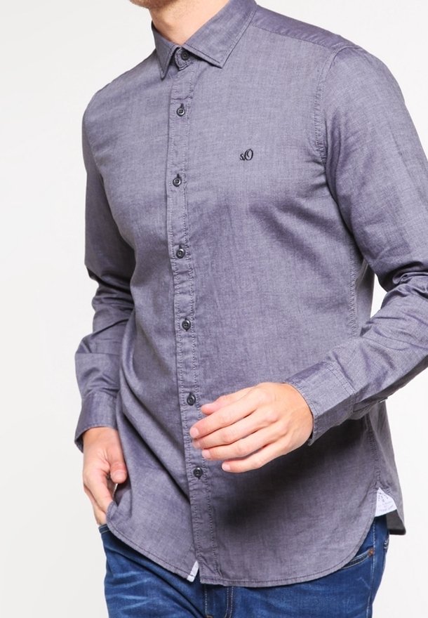 Homme portant une chemise grise à manches longues boutonnée avec un petit logo brodé sur la poitrine et un jean bleu.