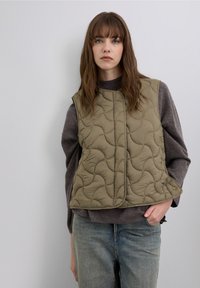 Quilted olive green vest with a smooth texture, featuring a round neckline and front snap closure, layered over a dark sweater.