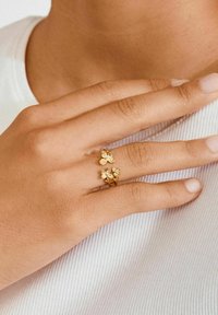 Kuldvärvid liblikamotiividega, millel on väikesed läbipaistvad kivid. Kandmisel käel, millel on loomulik küünelakk, tekstureeritud valge tausta ees.