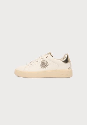Baskets basses beiges avec semelle crème, languette métallique dorée au talon, lacets et logo en forme de bouclier sur le côté sur fond blanc.