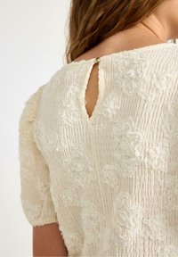 Kermanvärinen, kukkakuosisilla tekstiileillä varustettu pusero, jossa on kerrostettu muotoilu, lyhyet hihat ja avainlokki takana pienen nappikiinnityksen kanssa.