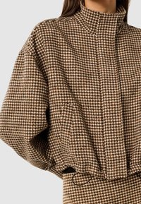Brun og beige houndstooth-mønster jakke med høj krave, løs pasform og samlede manchetter; lavet af tekstureret materiale.
