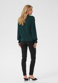 Dunkelgrüne Bluse mit langen Ärmeln und elastischen Bündchen, kombiniert mit schwarzen Slim-Fit-Jeans und schwarzen Absatzmules, die eine glatte Stofftextur präsentieren.