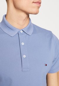 Light blue polo shirt made of textured fabric, featuring a collar, three buttons, and a small logo with red, white, and blue colors on the chest.