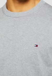 Primo piano di una persona che indossa un maglione grigio chiaro con scollatura a girocollo e un piccolo logo rettangolare rosso, bianco e blu sul petto.