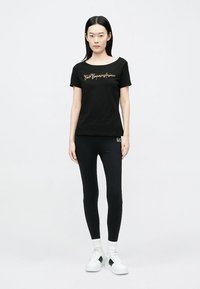 T-shirt nera in cotone con maniche corte e scollo ampio, con scritte dorate. Abbinata a leggings neri e scarpe da ginnastica bianche.