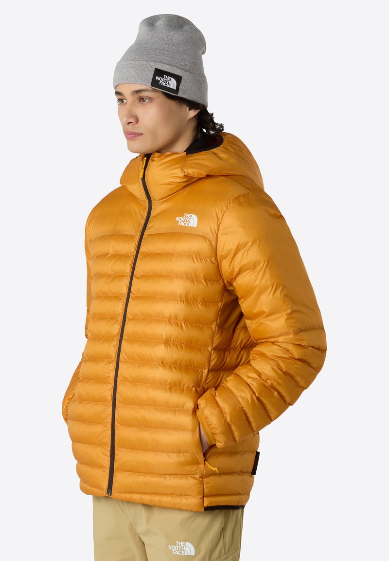 Bunda s náplní v jasně oranžové barvě s horizontálním prošíváním, vybavená kapucí, zipovým uzavřením a logem na prsíčku. Nositelná s šedou čepicí.