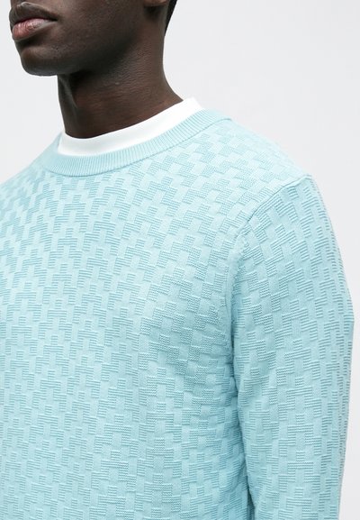 Pull en maille bleu clair avec un motif géométrique texturé, col rond et poignets côtelés. Aucun logo ou ornement visible.