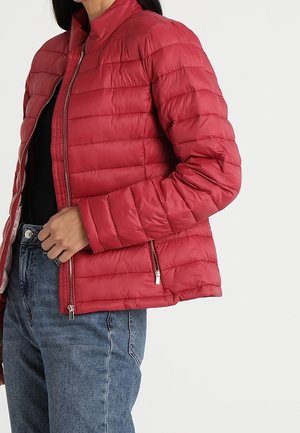 Red quilted puffer jacket with zipper pockets worn over a black top and blue jeans on a person against a plain background.
