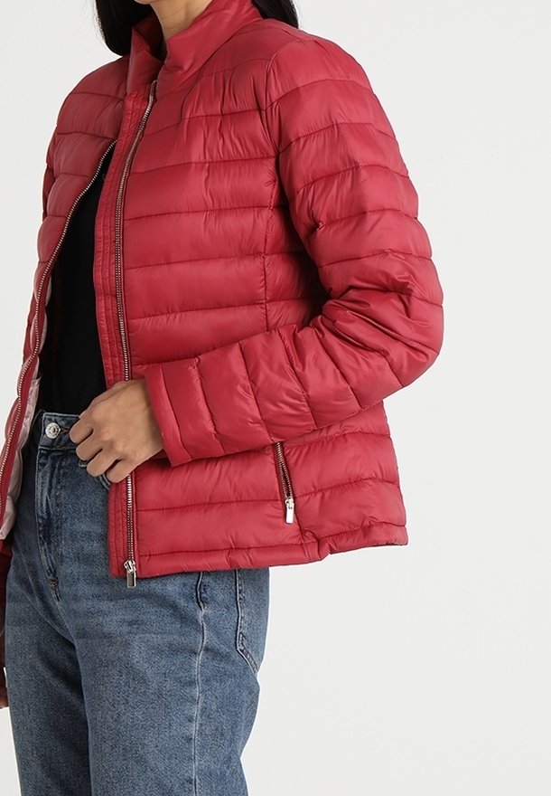 Doudoune matelassée rouge avec poches zippées portée sur un top noir et un jean bleu sur une personne devant un fond uni.