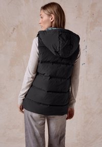 Schwarze Daunenweste mit Kapuze, ausgestattet mit horizontaler Steppung, Reißverschlusstaschen und einem Innenfutter in Grau. Getragen über einem hellgrauen Langarmoberteil.