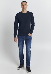 Pull en tricot bleu marine avec un col rond, associé à un jean en denim bleu clair et des baskets noires avec des semelles blanches. Design et coupe simples.