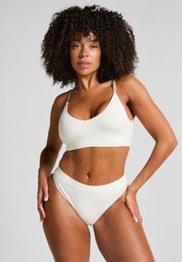 Vrouw met krullend haar in een witte geribbelde bralette en bijpassende hoge onderbroek, poserend met haar handen op haar heupen tegen een effen achtergrond.