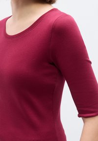Μπορντό μ πλούζα με μανίκια 3/4 από απαλό, υφασμάτινο ύφασμα με υφή· διαθέτει στρογγυλεμένο ντεκολτέ και εφαρμοστό σχέδιο που αναδεικνύει το σχήμα του σώματος.