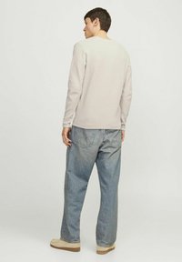 Hellbeige Langarmshirt aus strukturiertem Stoff; kombiniert mit weit geschnittenen blauen Jeans und hellbeigen Schuhen. Das Shirt hat einen runden Halsausschnitt.