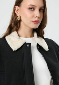 Zwarte jas met een crèmekleurige sherpa-kraag en rits. Voorzien van zilveren accenten en een eenvoudig, gestructureerd ontwerp. Draagt over een wit shirt.