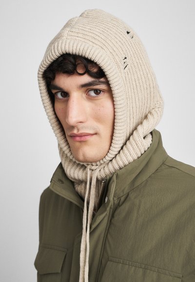 Bežena rebrasta pletena kapa s zategami, nosijoča se nad zelenim jopičem, ki ima tesno prileganje in majhne luknjice za teksturo.