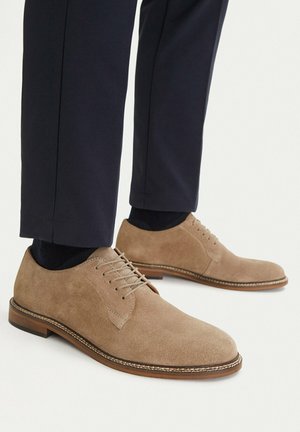 Beige suede lace-up dress shoes worn with black socks and dark navy trousers, shown from the side on a white background.