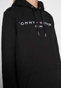 Schwarzer Hoodie aus weichem Baumwollmaterial; verfügt über eine Fronttasche, eine Kapuze mit Kordelzug und ein weißes gesticktes Logo mit roten Akzenten.