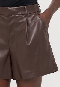 Shorts en cuir marron avec une texture lisse, un design plissé et des poches latérales. Coupés au-dessus du genou avec une coupe droite.