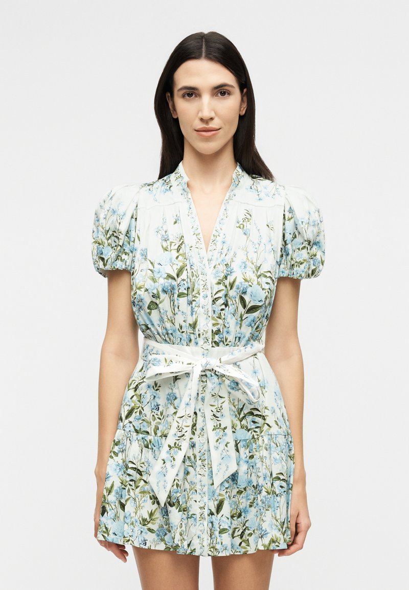 Woman wearing a white floral dress with puffed sleeves and a tied waist belt, standing against a plain light background.