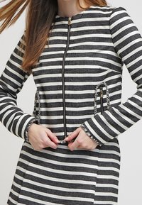 Czarna i biała kurtka w paski z okrągłym dekoltem, z zamkiem błyskawicznym z przodu oraz ozdobnymi suwakami na kieszeniach. Teksturowana tkanina.