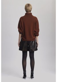 Rust-colored knitted sweater with a high collar, paired with shiny black leather shorts. Model wears black tights and high-heeled boots.