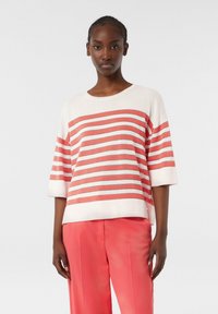 Gestreifter Strickpullover in Weiß und Koralle, mit weiten Ärmeln, lockerer Passform und geripptem Halsauschnitt. Kombiniert mit korallenfarbenen Hosen.