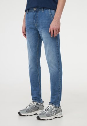 Jeans in denim blu chiaro con una vestibilità slim e un lavaggio sottile, dotati di zip frontale, passanti per cintura e tasche laterali. Abbinati a scarpe da ginnastica grigie.
