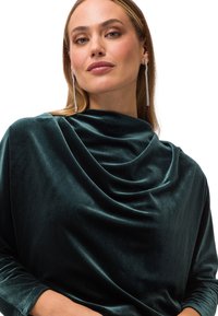 Grünes Samt-Oberteil mit Cowl-Neck-Design, langen Ärmeln und einer weichen Textur. Der Stoff hat einen glatten Glanz und fällt elegant.
