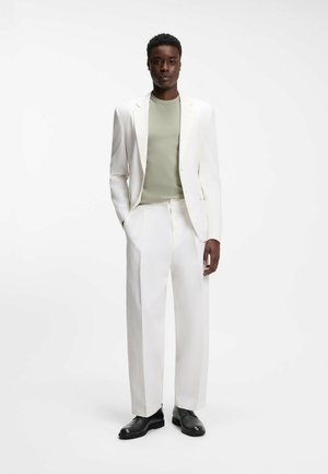 Uomo in piedi con completo bianco testurizzato, camicia verde chiaro e scarpe eleganti nere su sfondo bianco uniforme.