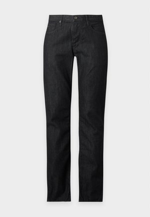 Homme debout portant un t-shirt blanc avec un logo noir, un jean foncé et des baskets noires sur un fond clair uni.