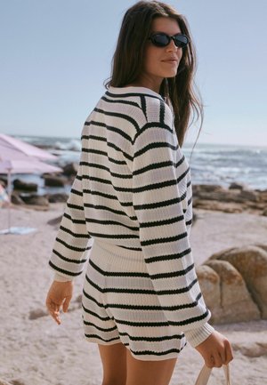 Frau trägt einen schwarz-weiß gestreiften Pullover und Shorts, Sonnenbrille, geht an einem felsigen Strand in Meeresnähe unter klarem Himmel spazieren.