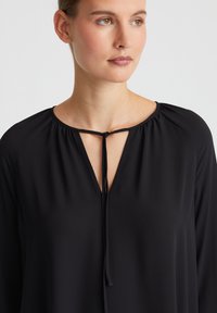 Schwarze Bluse mit V-Ausschnitt und Krawattenverschluss, langen Ärmeln und Raffung am Halsauschnitt, aus glattem, leichtem Stoff gefertigt.