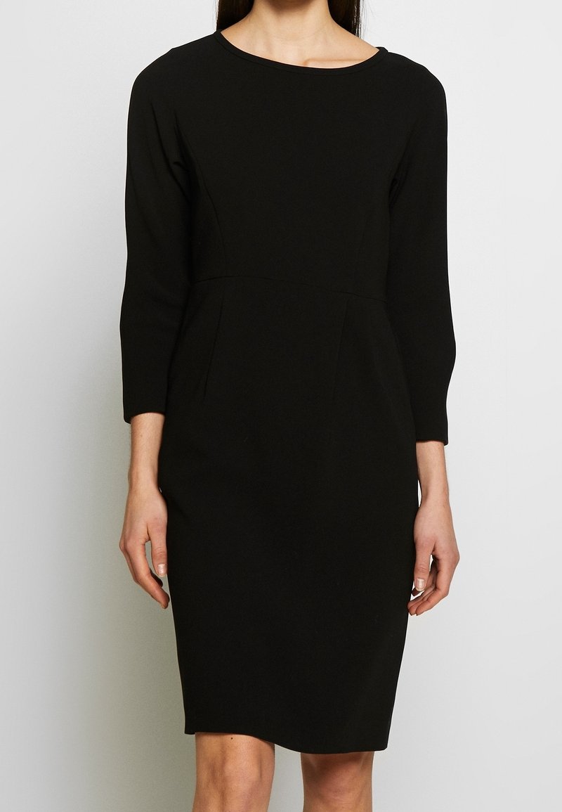 Femme portant une robe noire unie longueur genou avec des manches trois-quarts et un col rond, debout devant un fond clair uni.