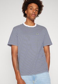 T-shirt rayé bleu marine et blanc avec un col rond blanc et des manches courtes. Présente un petit logo vert sur la poitrine. Matériau en coton.