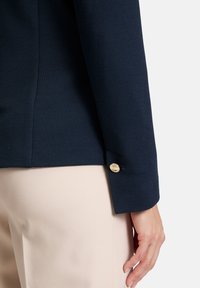 Granatowy żakiet o teksturowanym wykończeniu, charakteryzujący się strukturalnym krojem, złotymi guzikami oraz eleganckimi mankietami, zestawiony z jasnobeżowymi spodniami.